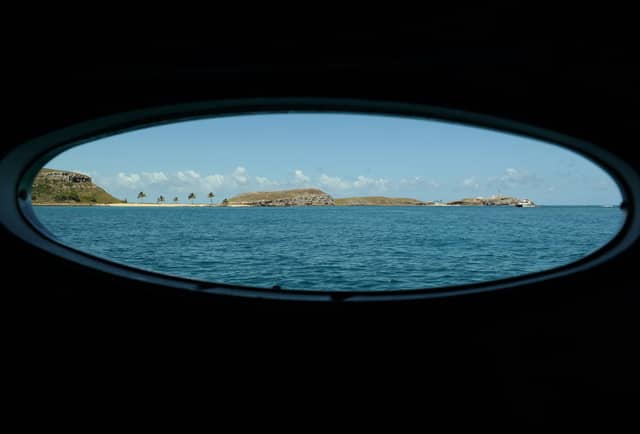 View through an oval boat porthole of the Abrolhos Islands rising from blue Atlantic waters, with palm trees, rocky hills, a lighthouse, and a moored vessel visible on the horizon during an abrolhos tour