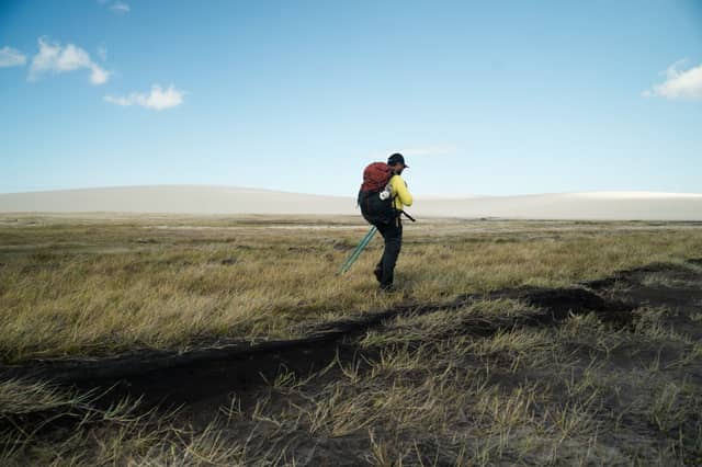 A solo hiker with a red backpack and trekking poles crosses open grassland during a hiking in Brazil adventure, with pale sand dunes rising in the distance under a wide blue sky.