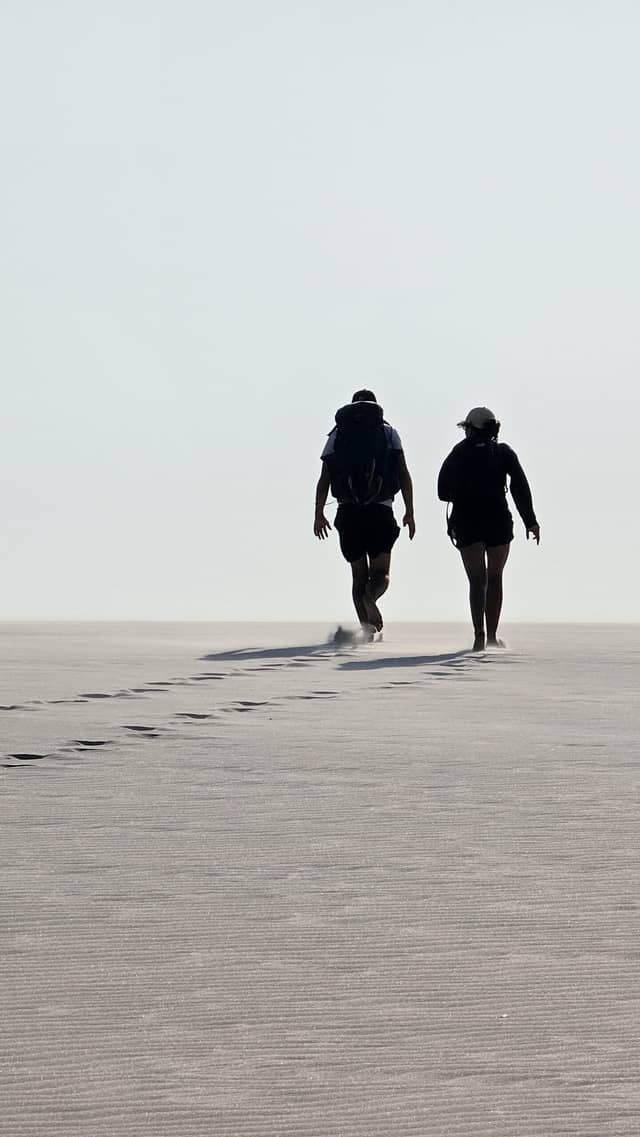 Two hikers with backpacks walking across a vast flat sand expanse during a hiking in brazil adventure, their footprints trailing behind them into the haze