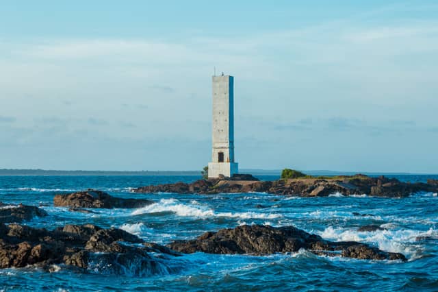 A tall concrete lighthouse standing on a rocky islet surrounded by churning blue Atlantic waves near Itacaré, Bahia, Brazil