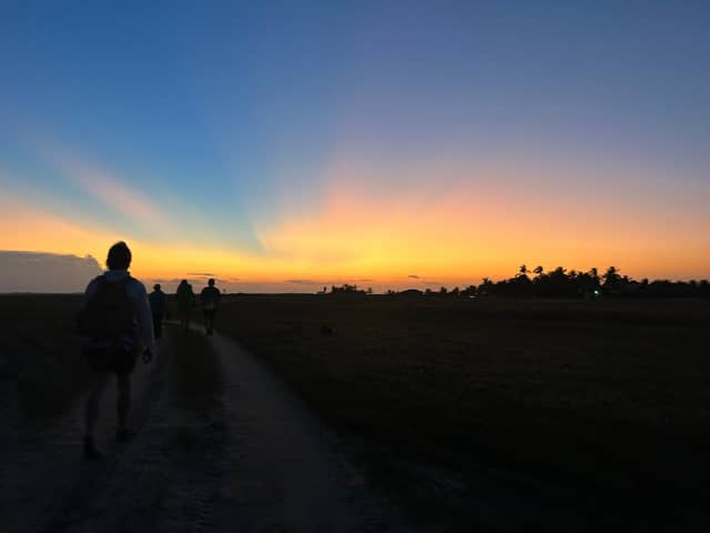 Silhouettes of trekkers walking a flat dirt trail toward a vivid orange and blue sunset, with palm trees on the horizon during trekking in Brazil
