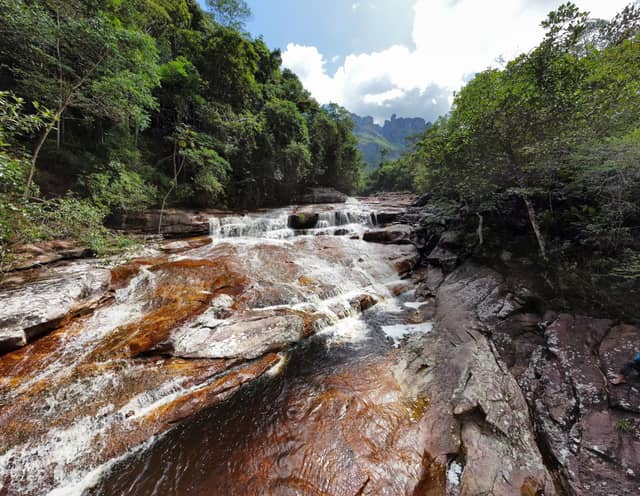 Shallow rapids cascade over iron-red sandstone slabs on the vale do pati trek, framed by dense Atlantic Forest and distant rocky peaks under a bright sky