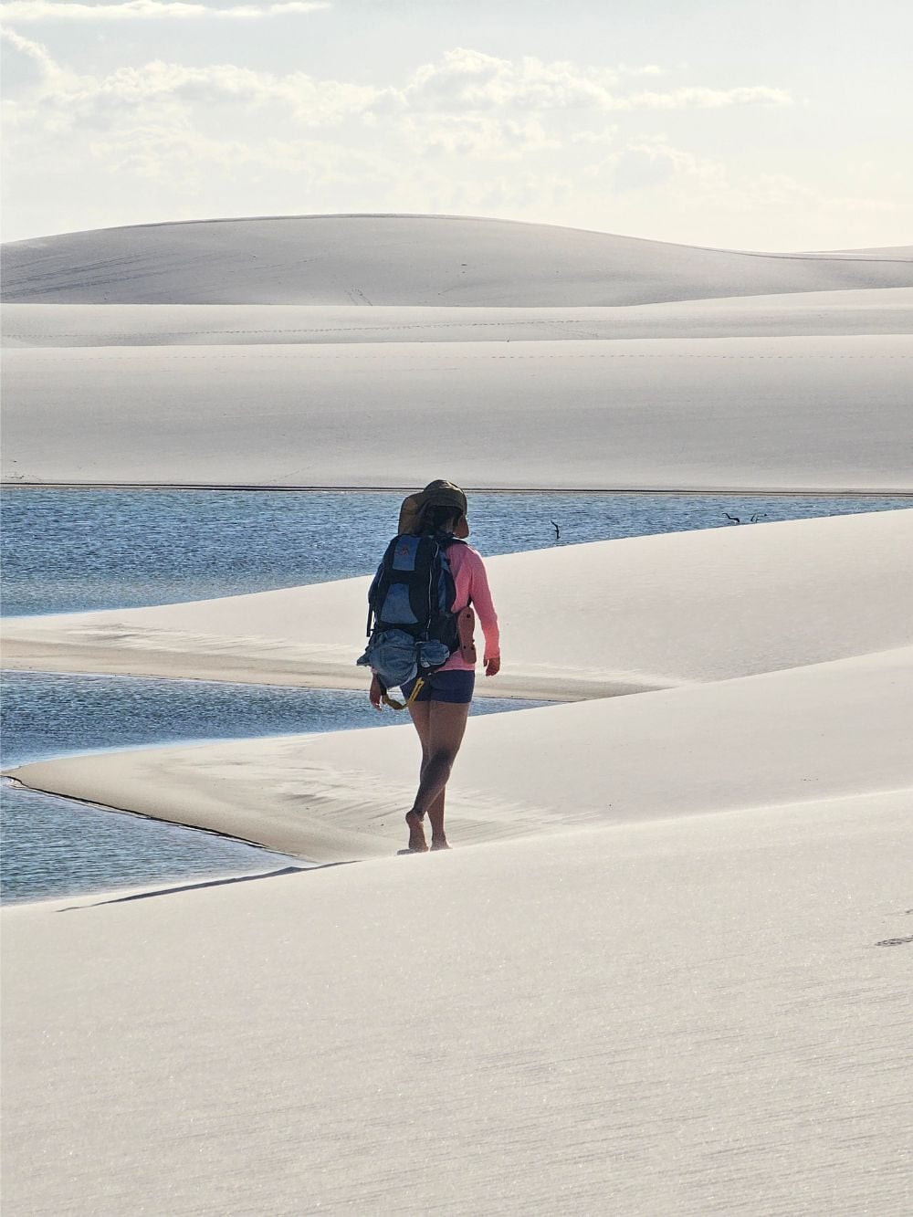 A backpacker walks barefoot along the white sand dunes near a blue rainwater lagoon while trekking in Lençóis Maranhenses National Park. 