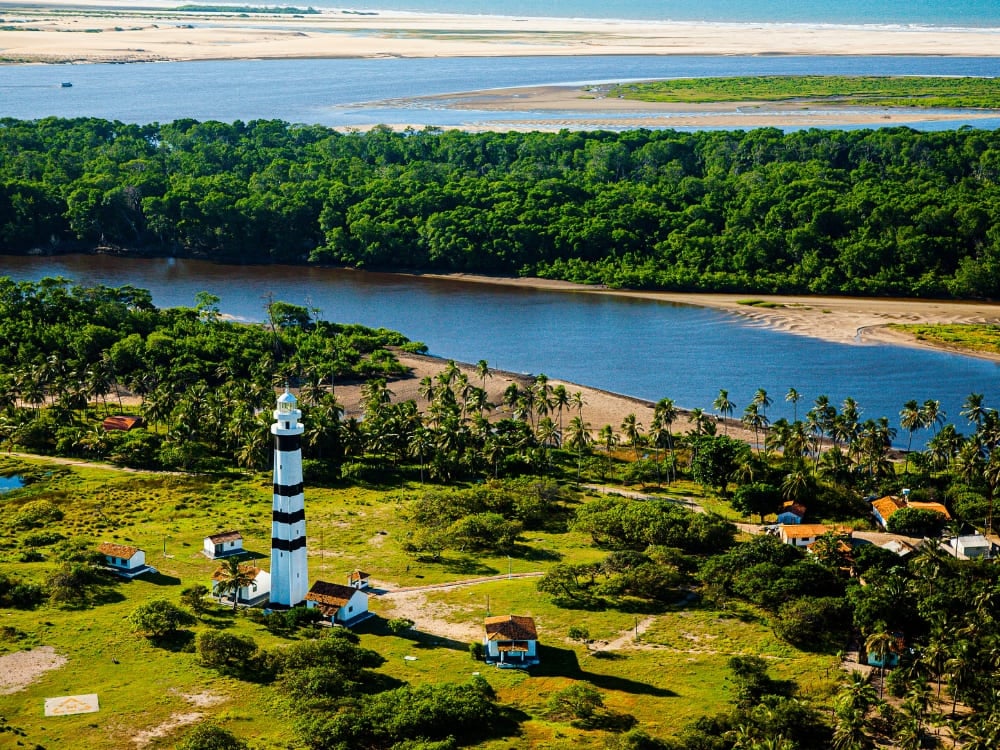 Aerial view of a tall black and white striped lighthouse and lush green vegetation near the base cities for Lençóis Maranhenses tours.