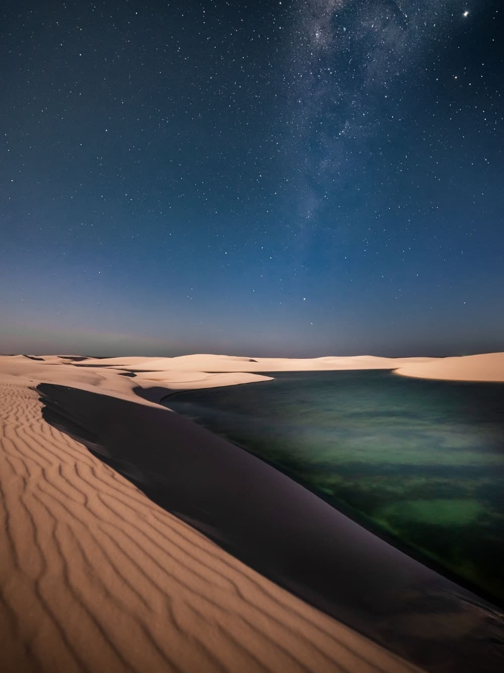 A serene nighttime landscape of sand dunes in Lençóis Maranhenses, Brazil, under a dark sky filled with stars, featuring a clear view of the Milky Way. In the foreground, a dark lagoon with hints of green and blue rests at the base of a rippled sand dune.