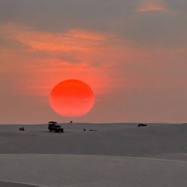 4x4 vehicles on the dunes under a large red setting sun in Lençóis Maranhenses