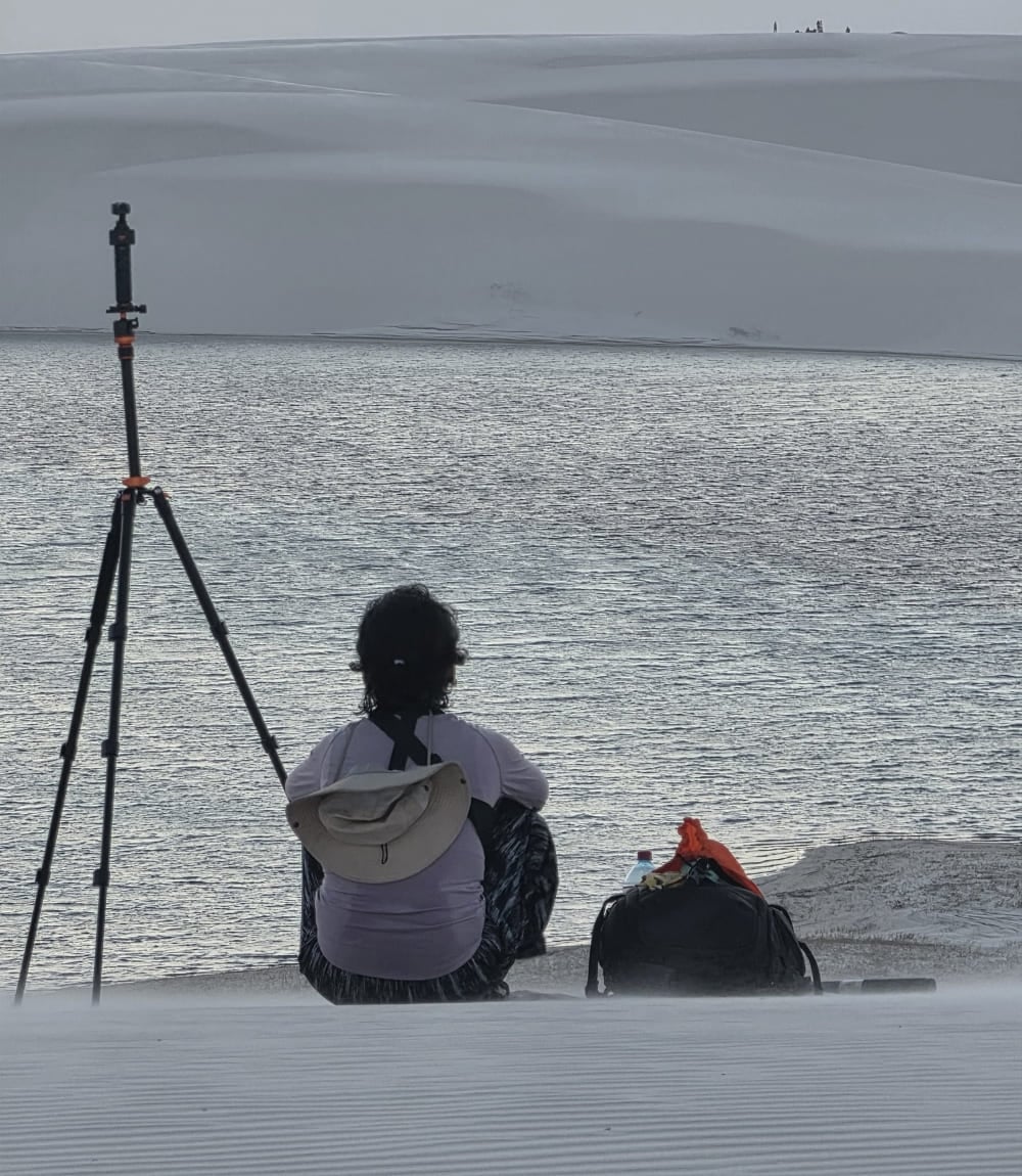 Person sitting on a dune beside a camera tripod, facing a lagoon in Lençóis Maranhenses