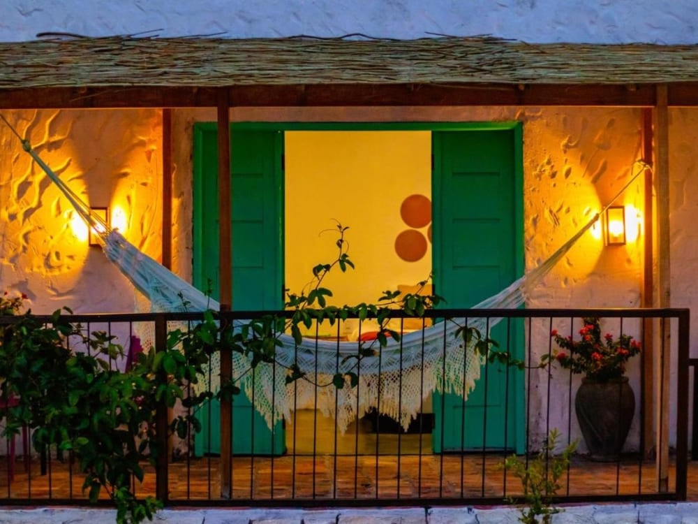 Intimate balcony lit at night with a resting hammock stretched in front of the green double door of a Quintal das Areias chalet. Intimate balcony lit at night with a resting hammock stretched in front of the green double door of a Quintal das Areias chalet.