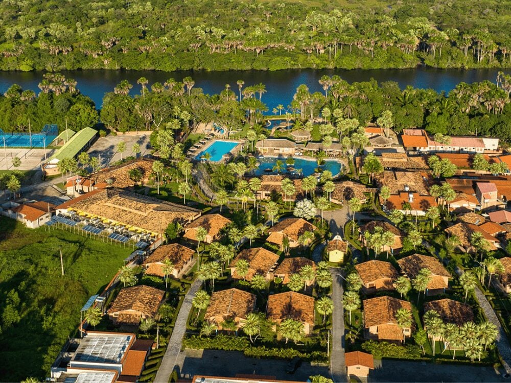 Photo: Porto Preguiça Resort Aerial view of a lush resort complex featuring red-roofed buildings and blue swimming pools nestled beside a wide river.