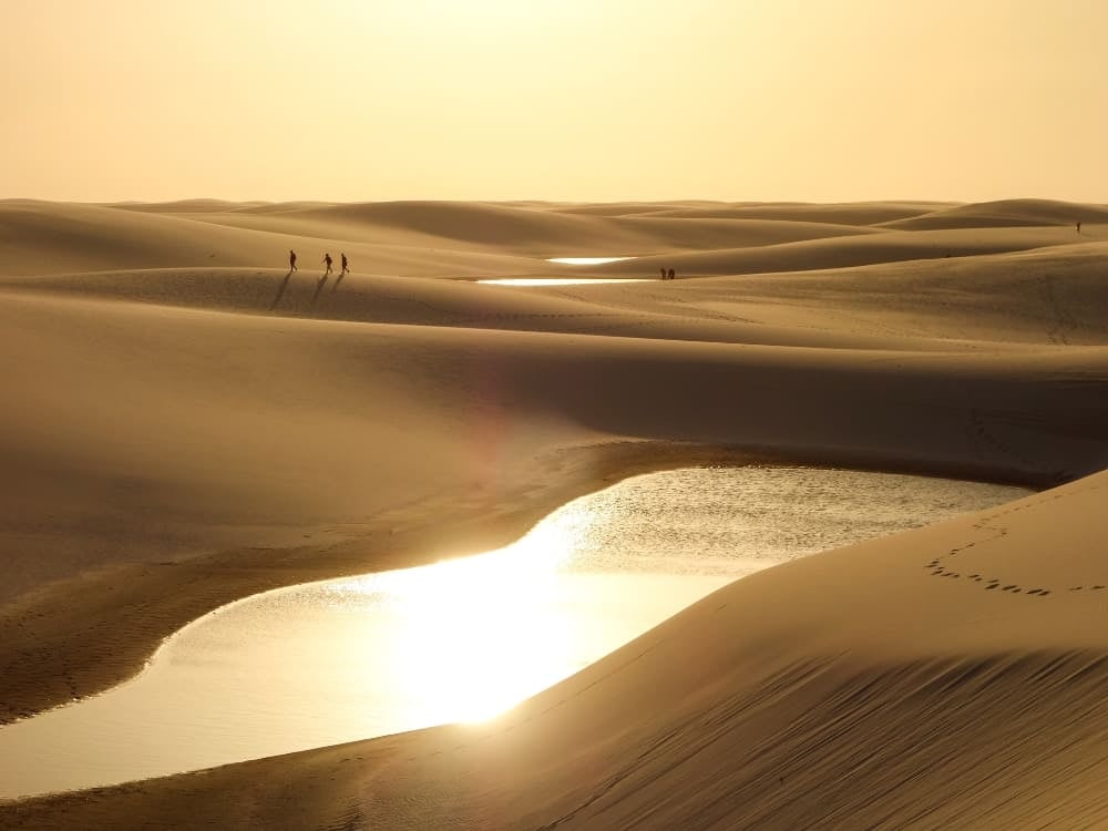 Sun setting over vast sand dunes with reflecting water pools while four people hike along the sandy ridge. Sun setting over vast sand dunes with reflecting water pools while four people hike along the sandy ridge.