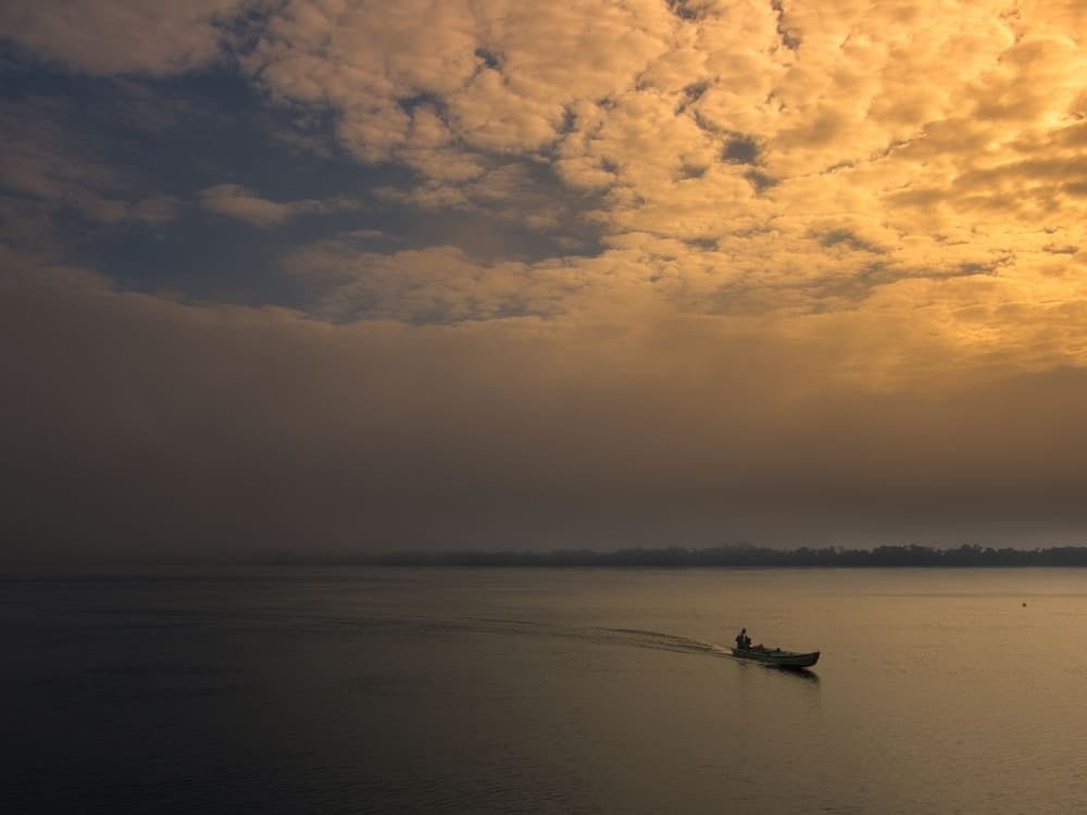A small motorized canoe traveling across the wide Amazon River during a golden sunrise under a cloudy sky. A small motorized canoe traveling across the wide Amazon River during a golden sunrise under a cloudy sky.