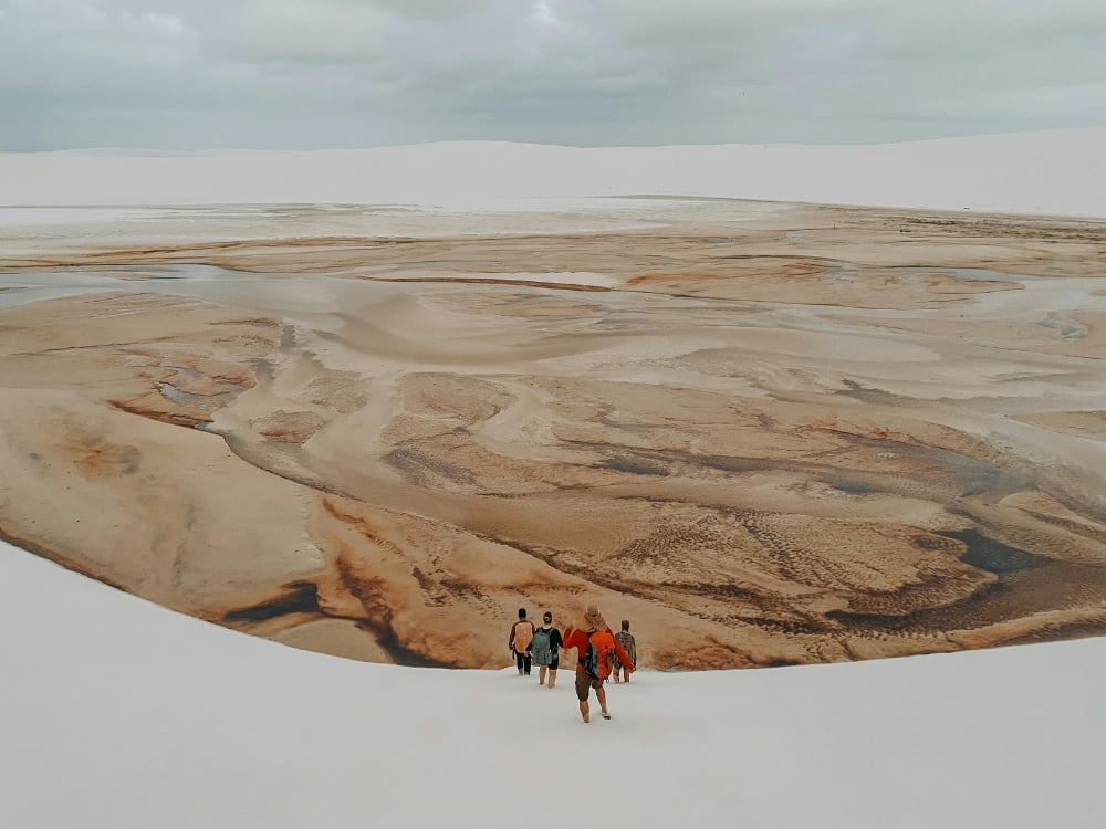 A group of tourists walks across a dry, brown lagoon bed surrounded by large white sand dunes during the dry season. A group of tourists walks across a dry, brown lagoon bed surrounded by large white sand dunes during the dry season.