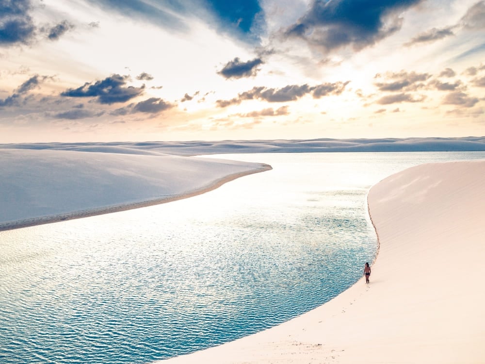 A traveler walking along the edge of a pristine blue lagoon surrounded by sand dunes during the best time to visit Lençóis Maranhenses A traveler walking along the edge of a pristine blue lagoon surrounded by sand dunes during the best time to visit Lençóis Maranhenses
