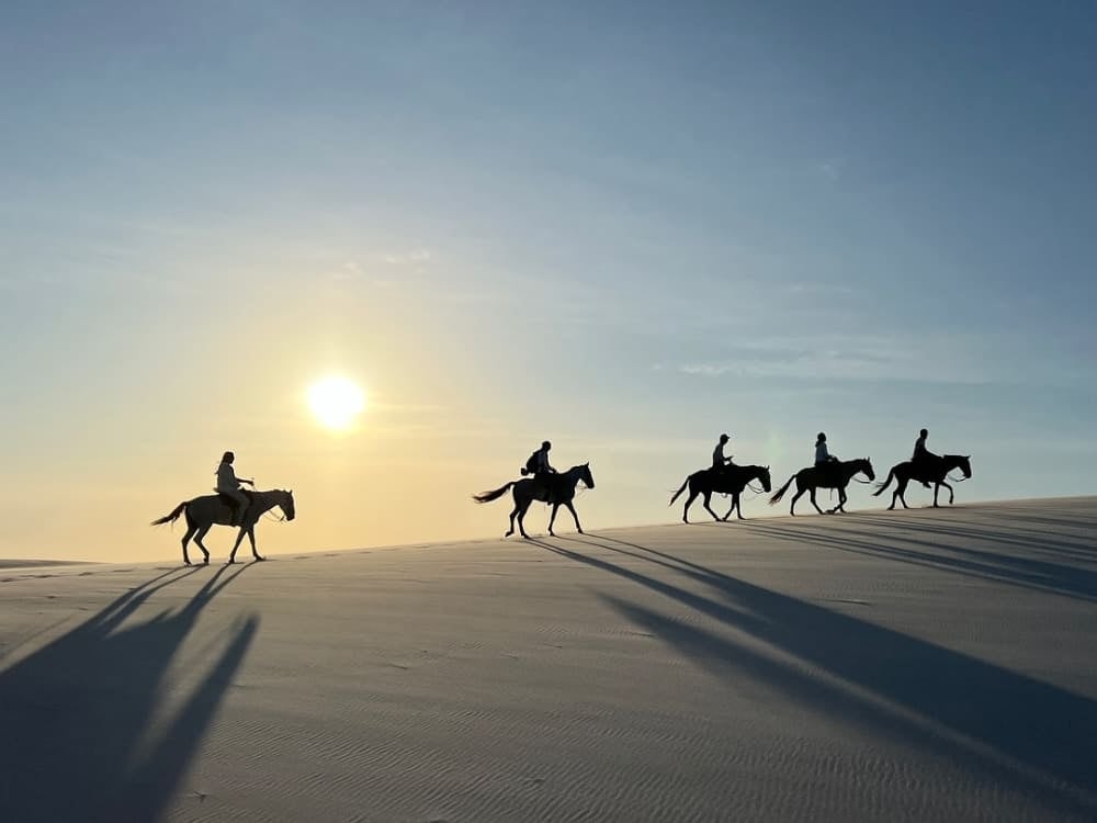 Silhouettes of five people horseback riding across the sand dunes of Lençóis Maranhenses under the bright late afternoon sun. Silhouettes of five people horseback riding across the sand dunes of Lençóis Maranhenses under the bright late afternoon sun.