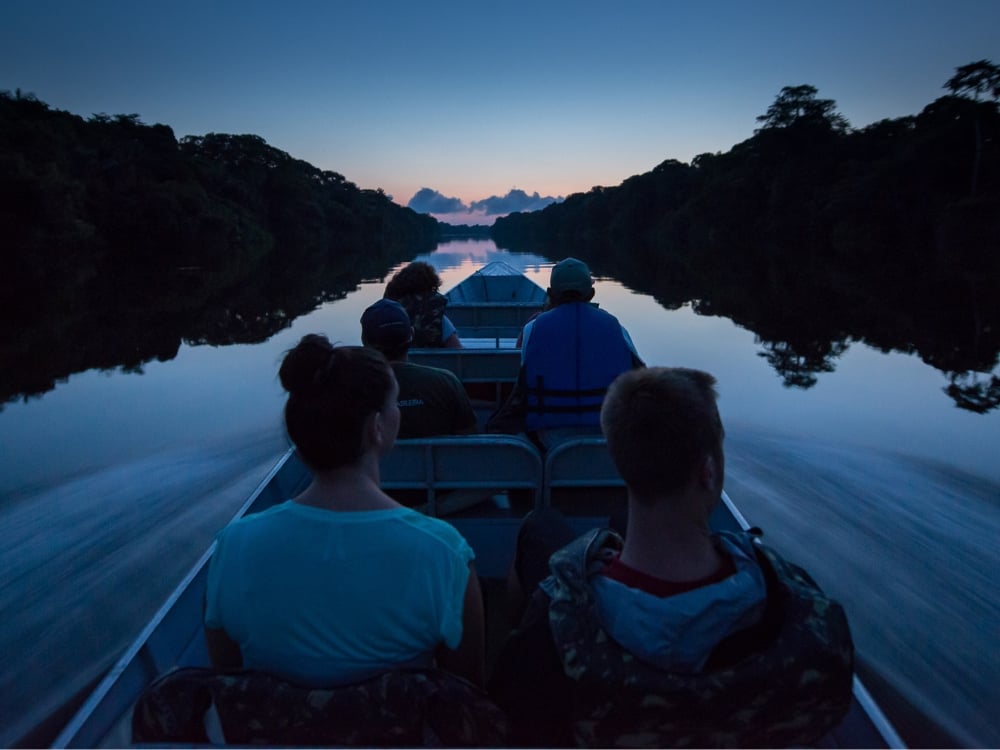 Tourists riding a motorized canoe exploring the calm waters of the Amazon Rainforest river at dusk Tourists riding a motorized canoe exploring the calm waters of the Amazon Rainforest river at dusk