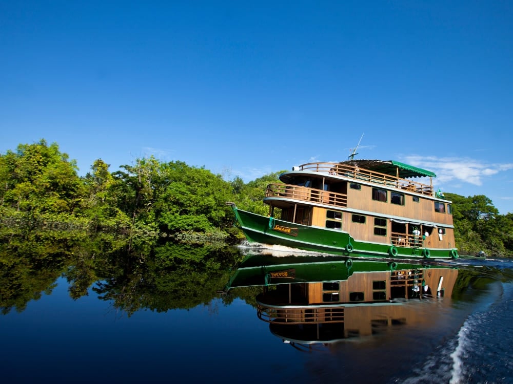 A rustic, three-deck wooden riverboat cruises smoothly along a dark Amazonian river beneath a blue sky. A rustic, three-deck wooden riverboat cruises smoothly along a dark Amazonian river beneath a blue sky.