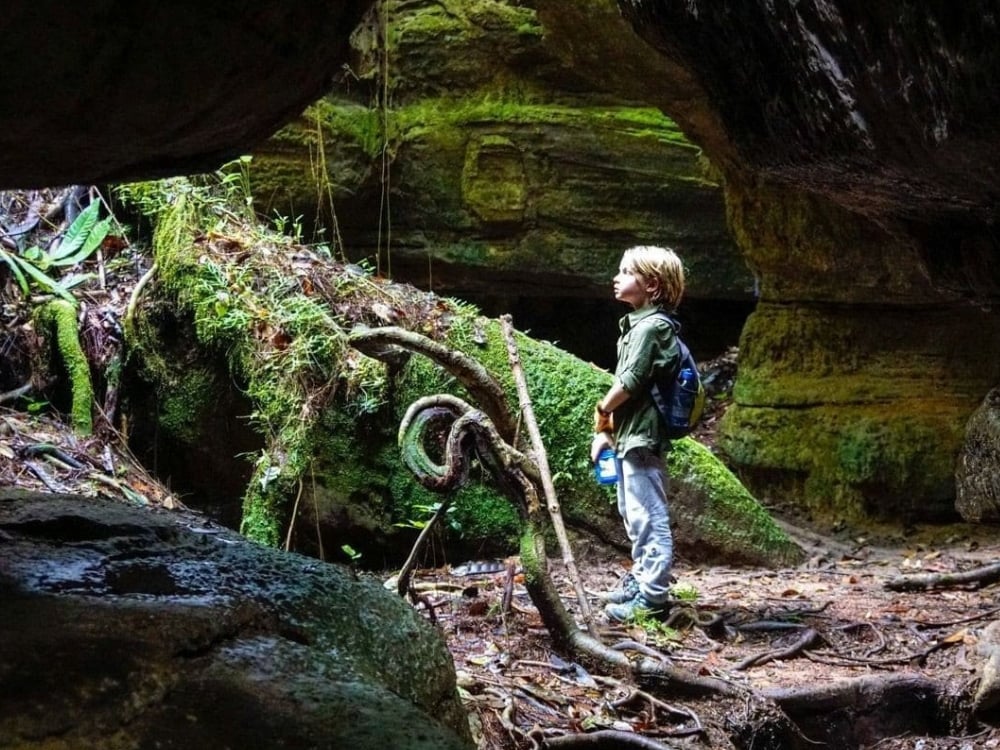 . Young boy standing at the entrance of a mossy cave exploring the Amazon jungle.