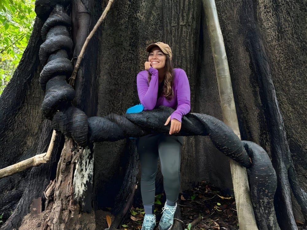 Smiling woman wearing a purple shirt, leaning against the thick, twisted roots of a giant tree in the middle of a lush green forest. Smiling woman wearing a purple shirt, leaning against the thick, twisted roots of a giant tree in the middle of a lush green forest.