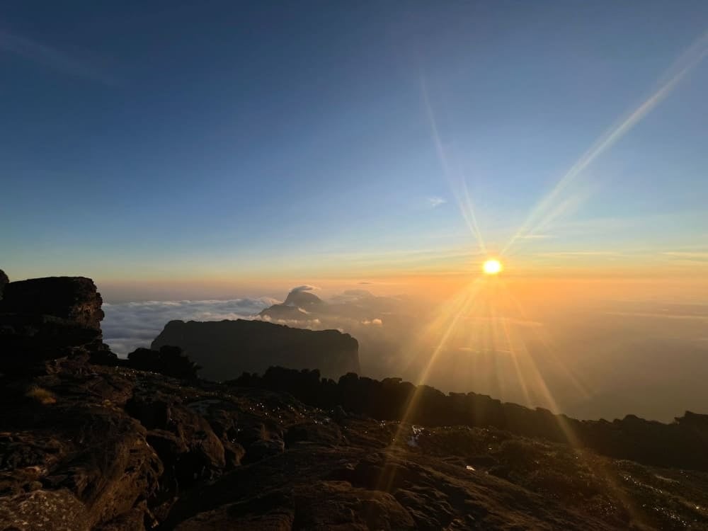 The sun bursts with bright rays over a sea of white clouds and silhouetted table mountains, viewed from the rocky summit of Mount Roraima. The sun bursts with bright rays over a sea of white clouds and silhouetted table mountains, viewed from the rocky summit of Mount Roraima.
