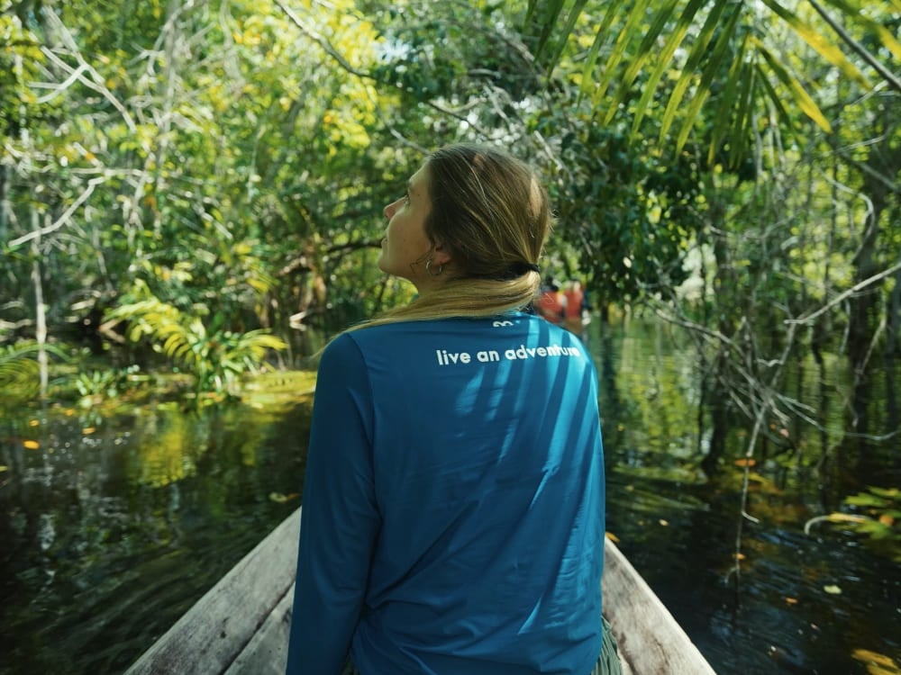 Woman from behind in a wooden boat navigating a river surrounded by dense forest, wearing a blue shirt with the phrase live an adventure. Woman from behind in a wooden boat navigating a river surrounded by dense forest, wearing a blue shirt with the phrase live an adventure.