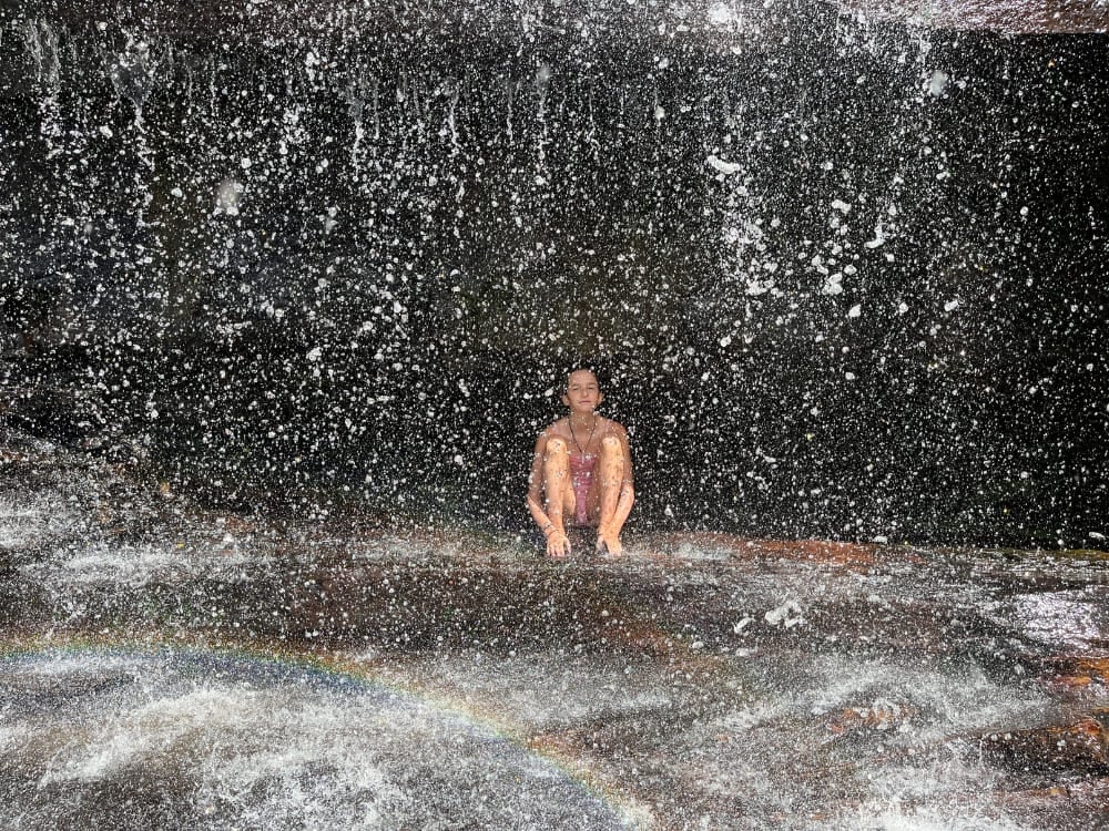 A person sits against a dark rock wall, enjoying a refreshing shower under the cascading cold waters of a mountain-fed waterfall. A person sits against a dark rock wall, enjoying a refreshing shower under the cascading cold waters of a mountain-fed waterfall.