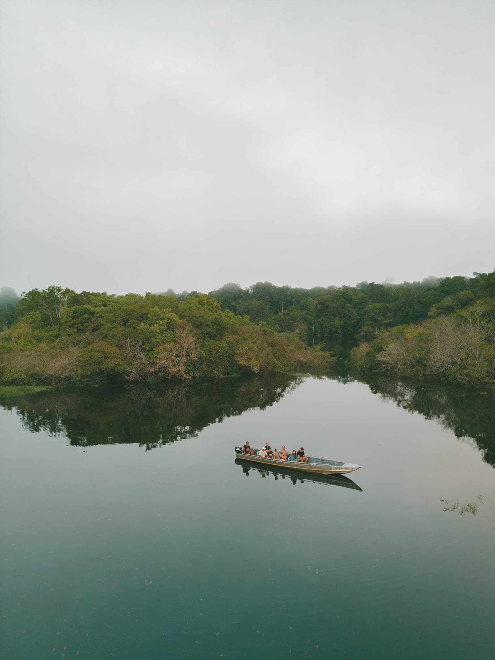 Six passengers, one river, and the forest holding its breath around them. A small motorboat carrying a group of travellers glides across a glassy, mist-softened river on an Amazon boutique lodge excursion, surrounded by dense tropical forest reflected in the still water