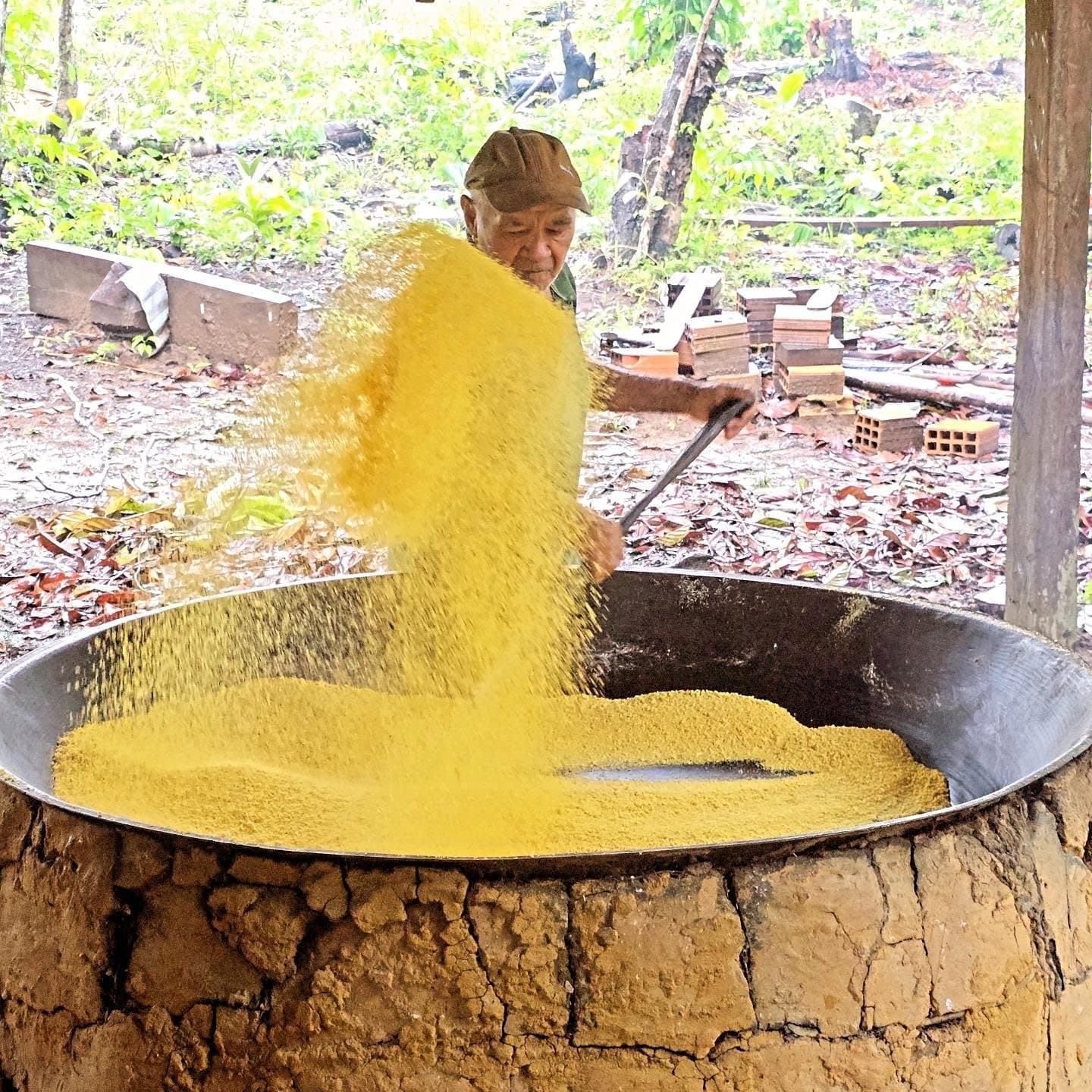 Farinha feita assim tem cheiro, tem história, e fica muito melhor no prato. An elder man toasts golden cassava flour in a large iron pan over a clay wood-fired oven during a community visit at an Amazon boutique lodge experience