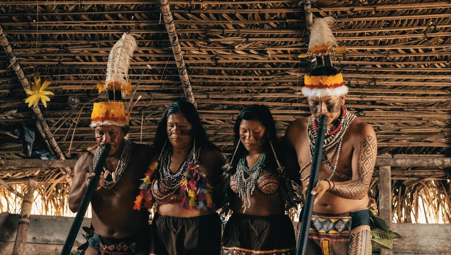 Songs older than any map, still very much alive. Four members of an Upper Rio Negro indigenous community in traditional dress, wearing feathered headdresses, bead necklaces, geometric body paint, and colorful feather ornaments, performing inside a thatched-roof longhouse during an Amazon Cruise Trip in Brazil