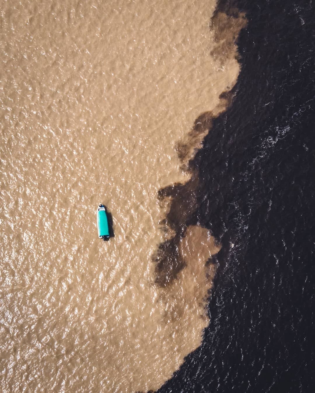 Two rivers, one boat, and a boundary that refuses to blur. Aerial view of a small teal-roofed river boat floating at the Meeting of the Waters, where brown sediment-laden water meets dark black-water river on this Amazon cruise trip in Brazil