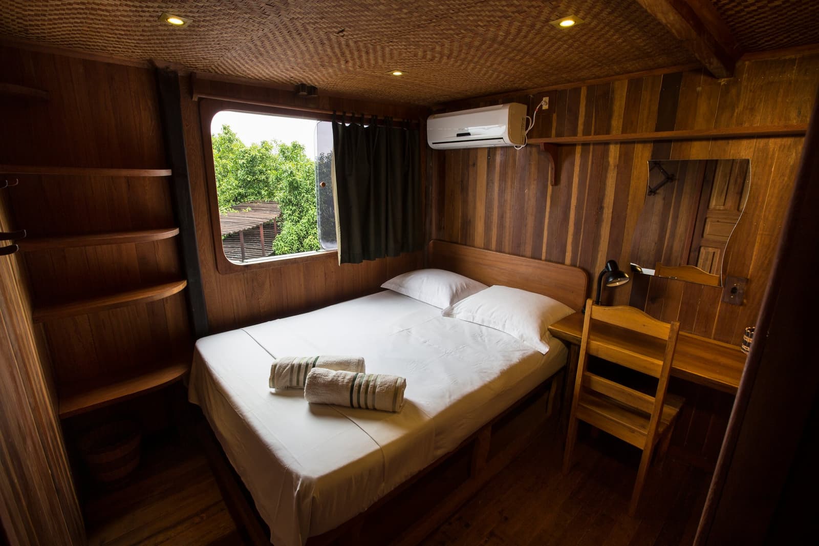 Small space, done right, wood, white linen, and the Amazon just outside the porthole. Wood-paneled private cabin on an Amazon Cruise Trip in Brazil, with a double bed dressed in white linen, two rolled towels, open shelves, a desk with a reading lamp, air conditioning, and a porthole-style window overlooking green rainforest canopy