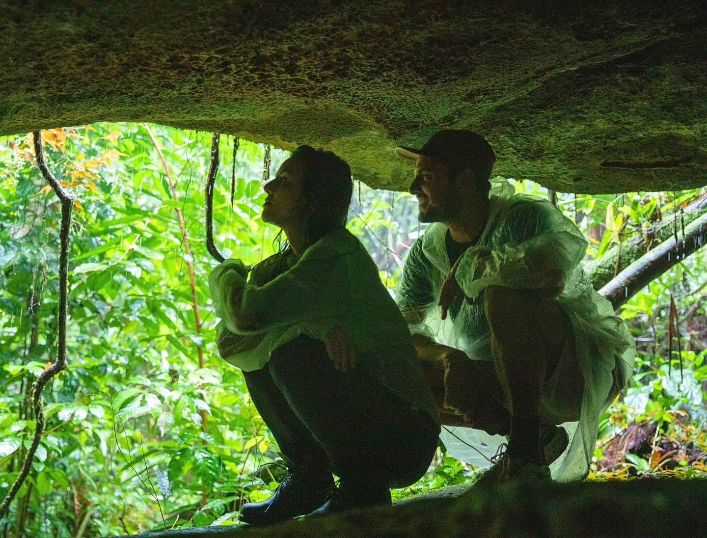 Rain mid-hike is not a setback. It is just the forest setting the pace. Two travelers crouch beneath a mossy rock overhang on an amazon river cruise brazil forest hike, surrounded by dense green jungle foliage while sheltering from rain in their ponchos