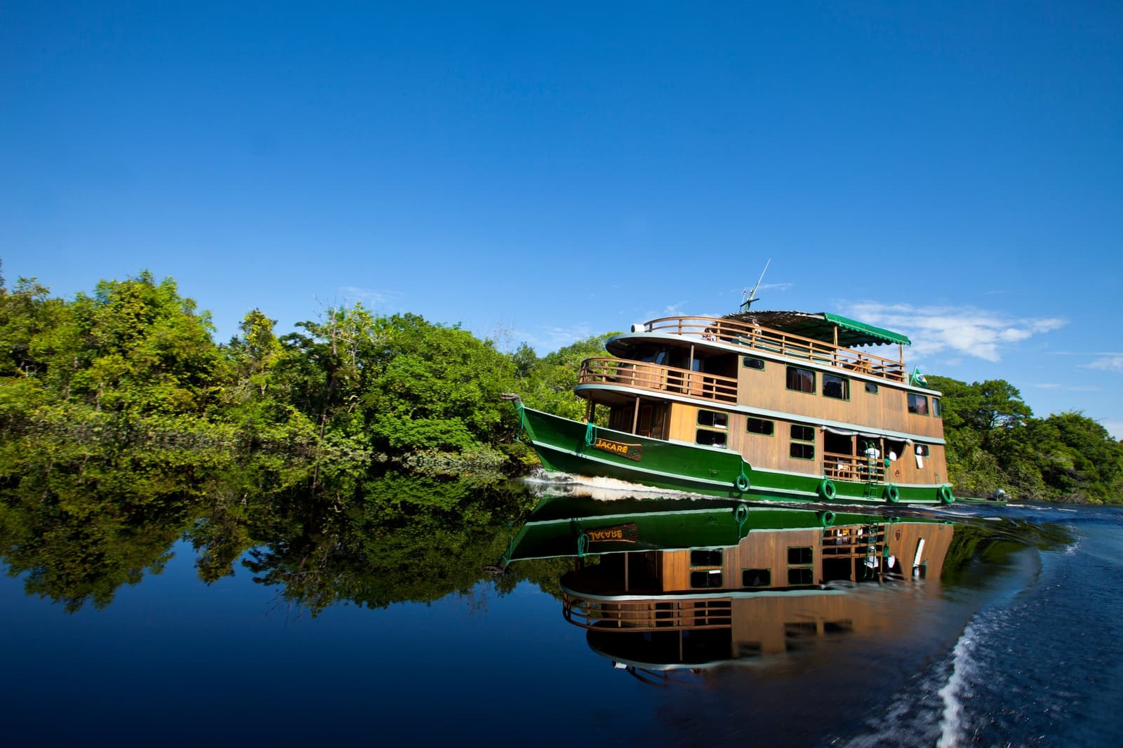 Home for five nights, and she handles the jungle like she owns it. The Jacaré riverboat, a three-deck wooden vessel with green hull, cruising a dark calm river on an amazon river cruise in Brazil, with dense forest lining both banks under a clear blue sky