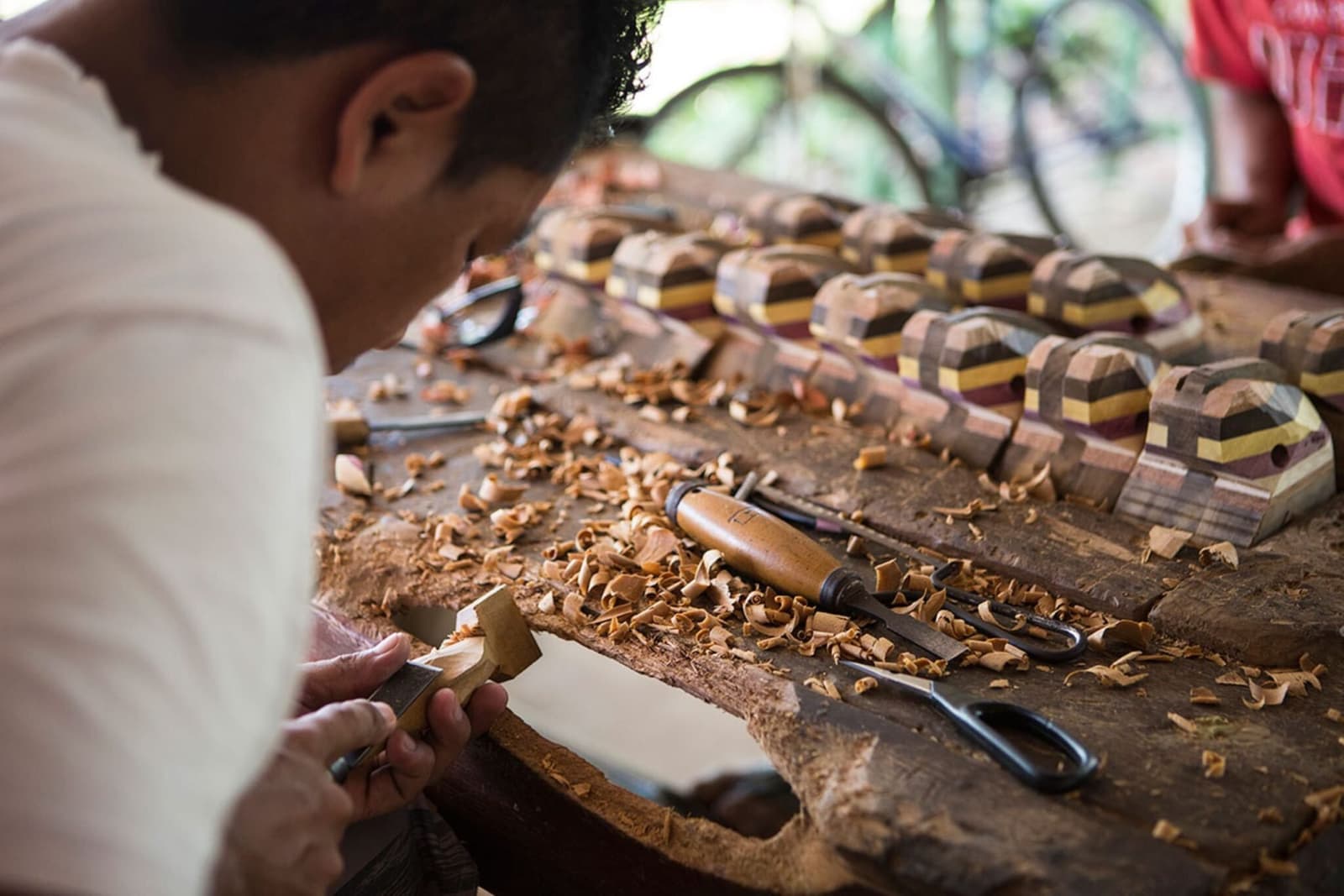Craft this good takes years of practice, and about five minutes to fall in love with. A craftsman carves a small wooden piece by hand at a workbench covered in wood shavings, with rows of multicolored laminated wood figurines lined up beside his tools during a stop on an amazon river cruise brazil itinerary