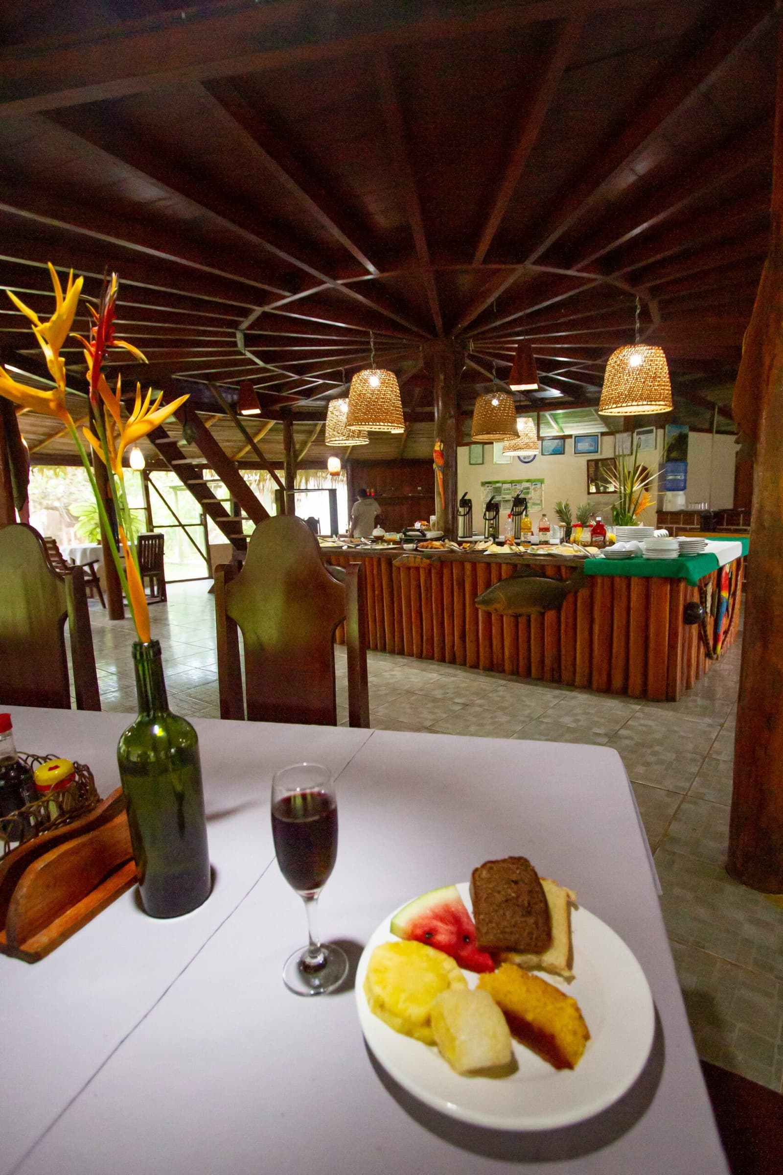 Breakfast sorted before the forest even wakes up. The Amazon Turtle Lodge dining room features a spread of fresh tropical fruit, bread, and red wine on a foreground table, with a full buffet counter, woven pendant lights, and a dramatic hardwood ceiling visible beyond