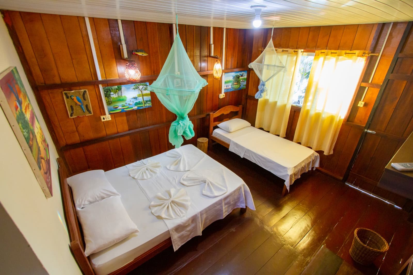 Cosy enough to forget you just hiked through the rainforest. A timber-panelled twin room at Amazon Turtle Lodge with a double bed and single bed, green and white mosquito nets hanging overhead, folded towel art on white linens, and colourful rainforest paintings on the walls