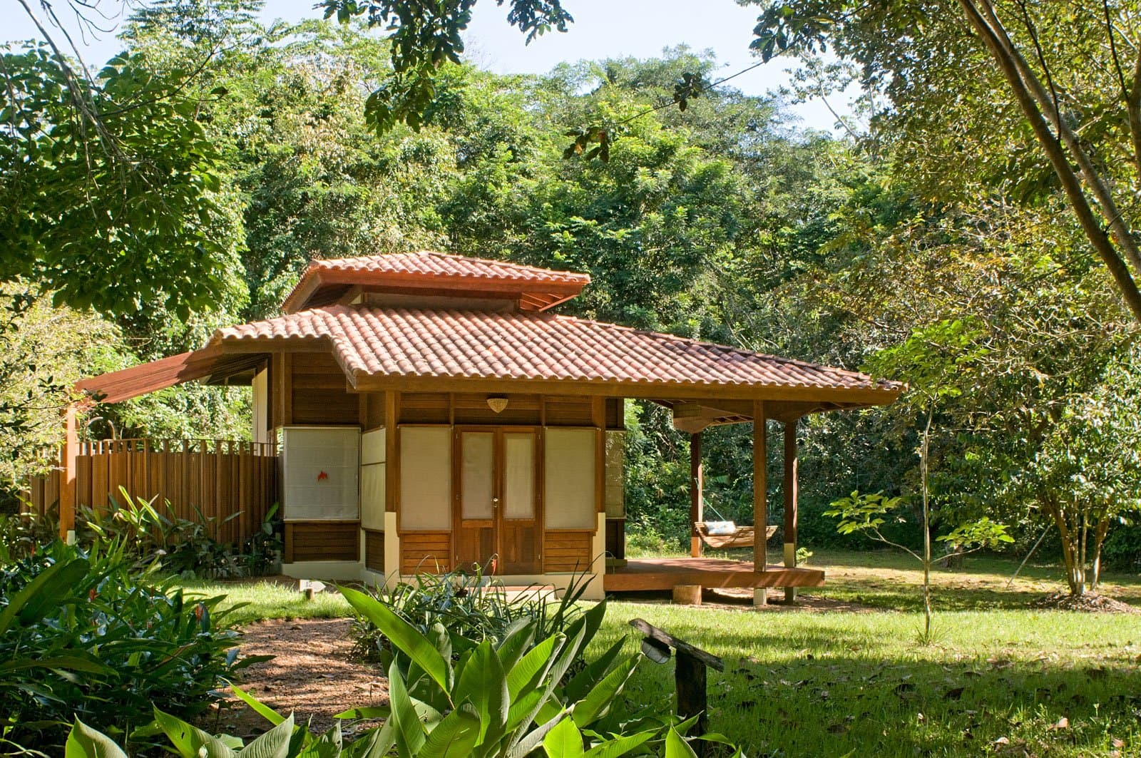 Your base between dawn birding walks: a shaded veranda and a hammock that earns its keep. Photo: Luis Gomes A wooden bungalow with a terracotta-tiled roof and covered veranda with a hammock, set among dense Amazon rainforest trees — accommodation for birdwatching in the Amazon rainforest at Cristalino Jungle Lodge