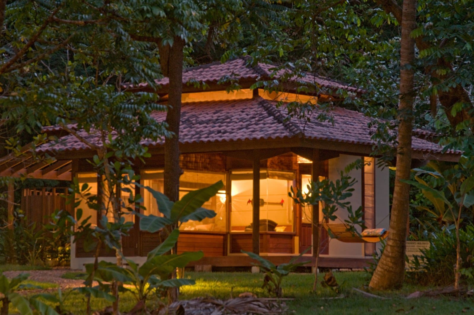 Warm light, forest closing in, hammock waiting. Hard to leave in the morning. Photo: Luis Gomes A warmly lit wooden bungalow with tiled roof and glass walls glowing amber at dusk, surrounded by dense Amazon rainforest trees and tropical foliage, with a hammock on the veranda — ideal base for birdwatching in the Amazon rainforest