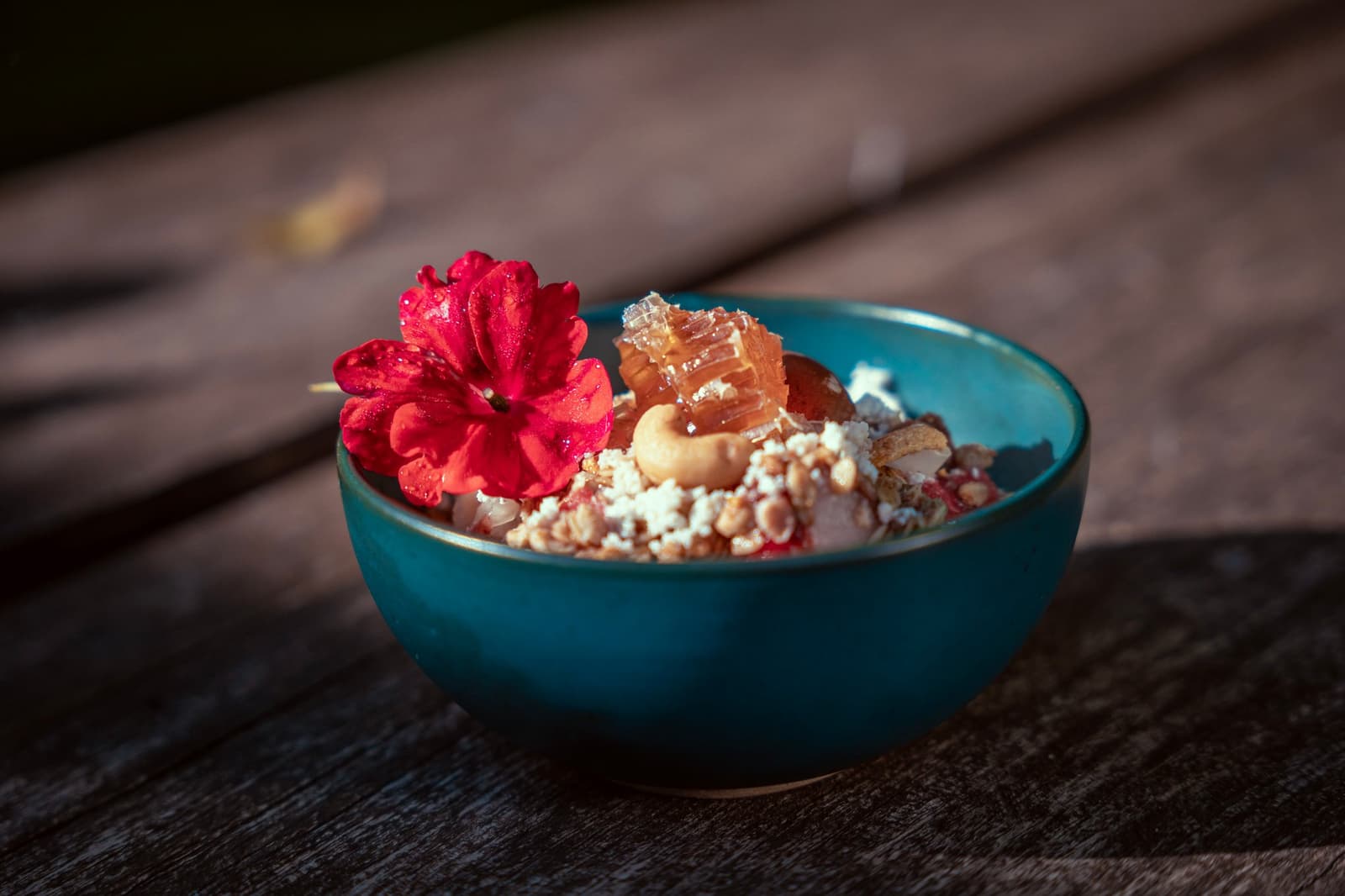 Breakfast here earns its flower. Fuel up before the birds do. A teal ceramic bowl filled with granola, cashews, honeycomb, and crumbled cheese, garnished with a red flower, served on a rustic wooden table at a birdwatching in the amazon rainforest lodge