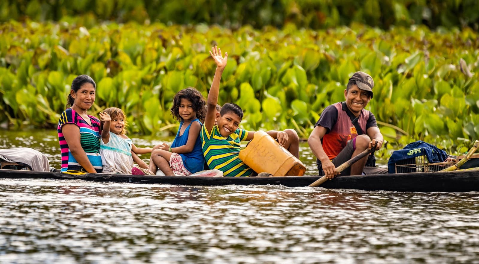 The best hello you'll get all trip, guaranteed. A smiling family of five paddles a wooden dugout canoe through a river on a brazilian amazon jungle tour, with a boy waving enthusiastically and lush aquatic vegetation in the background