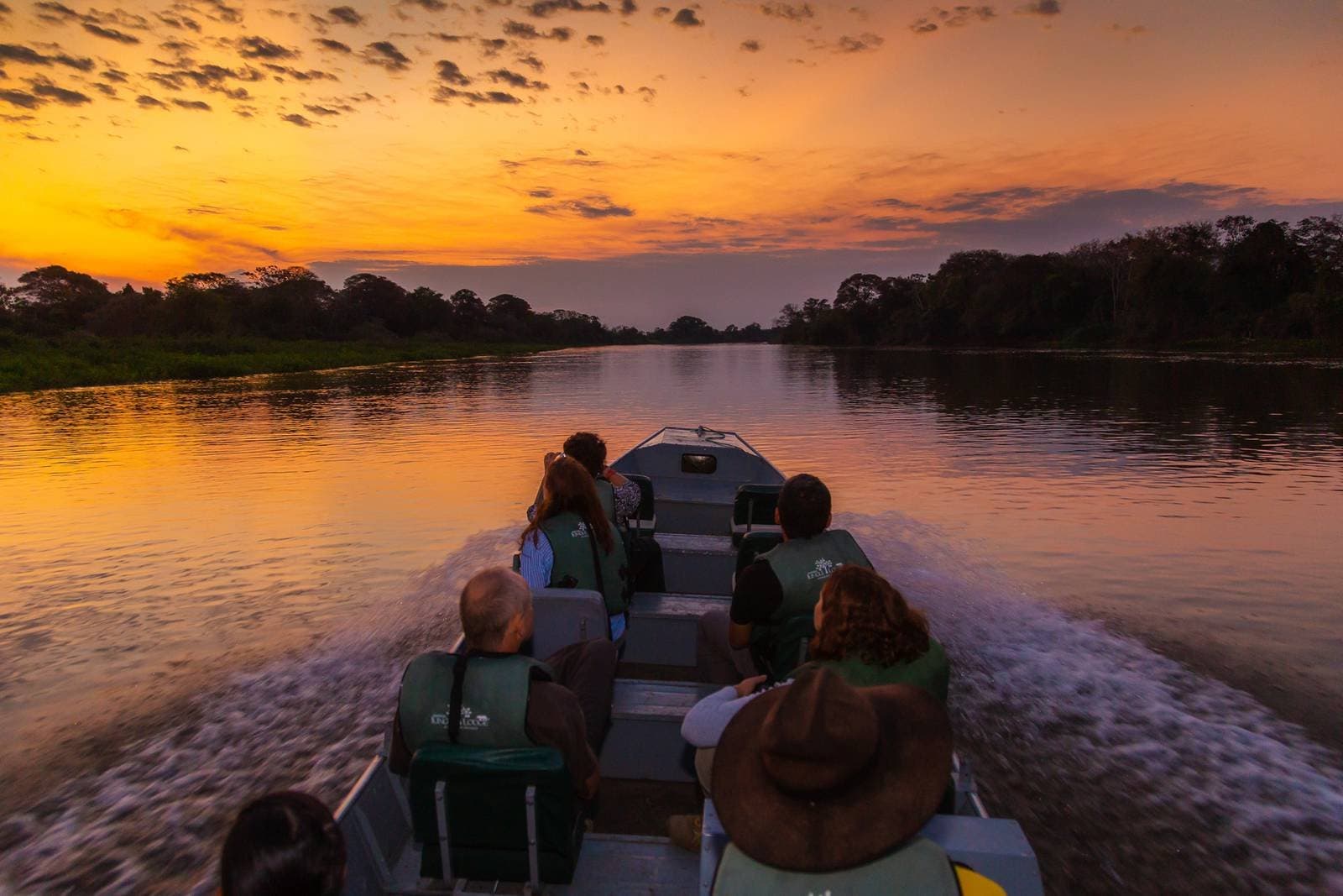 The best seat in the house, and it floats. Travelers in life jackets on a motorboat cruising a calm river channel during a vivid orange and gold sunset on a budget Pantanal tour, with dense forest silhouetted on both banks