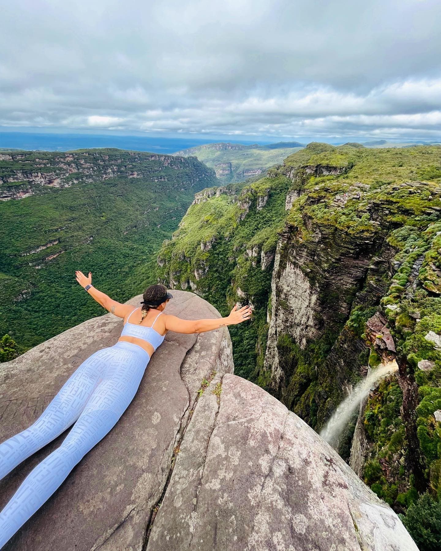 Arms wide open, canyon dropping away below, some views earn the nerves. A woman lies arms-wide on a boulder at the edge of a deep canyon on the Chapada Diamantina Tour, with a waterfall cascading down the cliff face and green forested plateaus stretching to the horizon