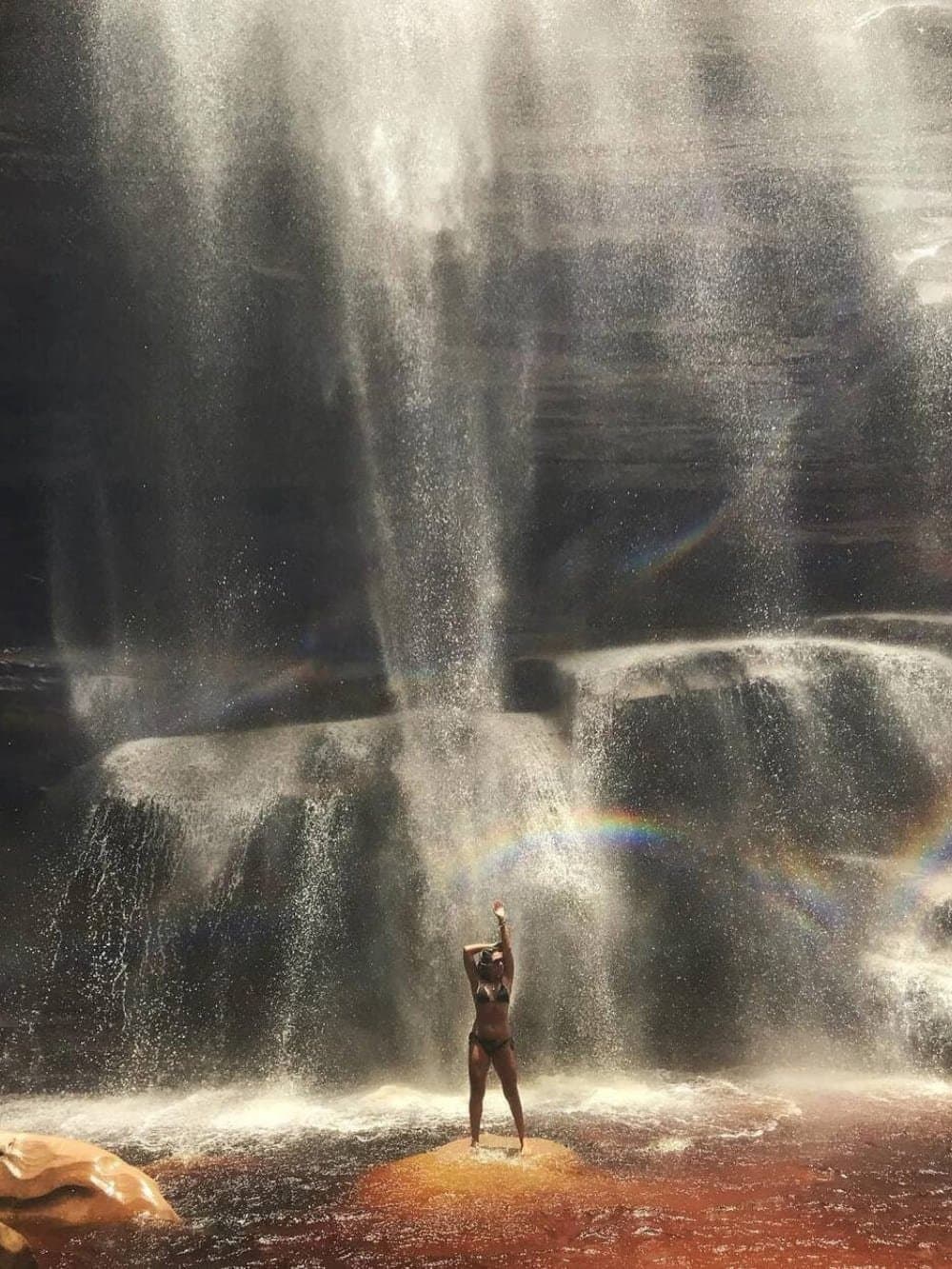 A rainbow you didn't have to chase, it came to you. A visitor in a bikini stands arms raised on a rock beneath a large waterfall on the Chapada Diamantina Tour, with a vivid rainbow arcing through the spray in the foreground