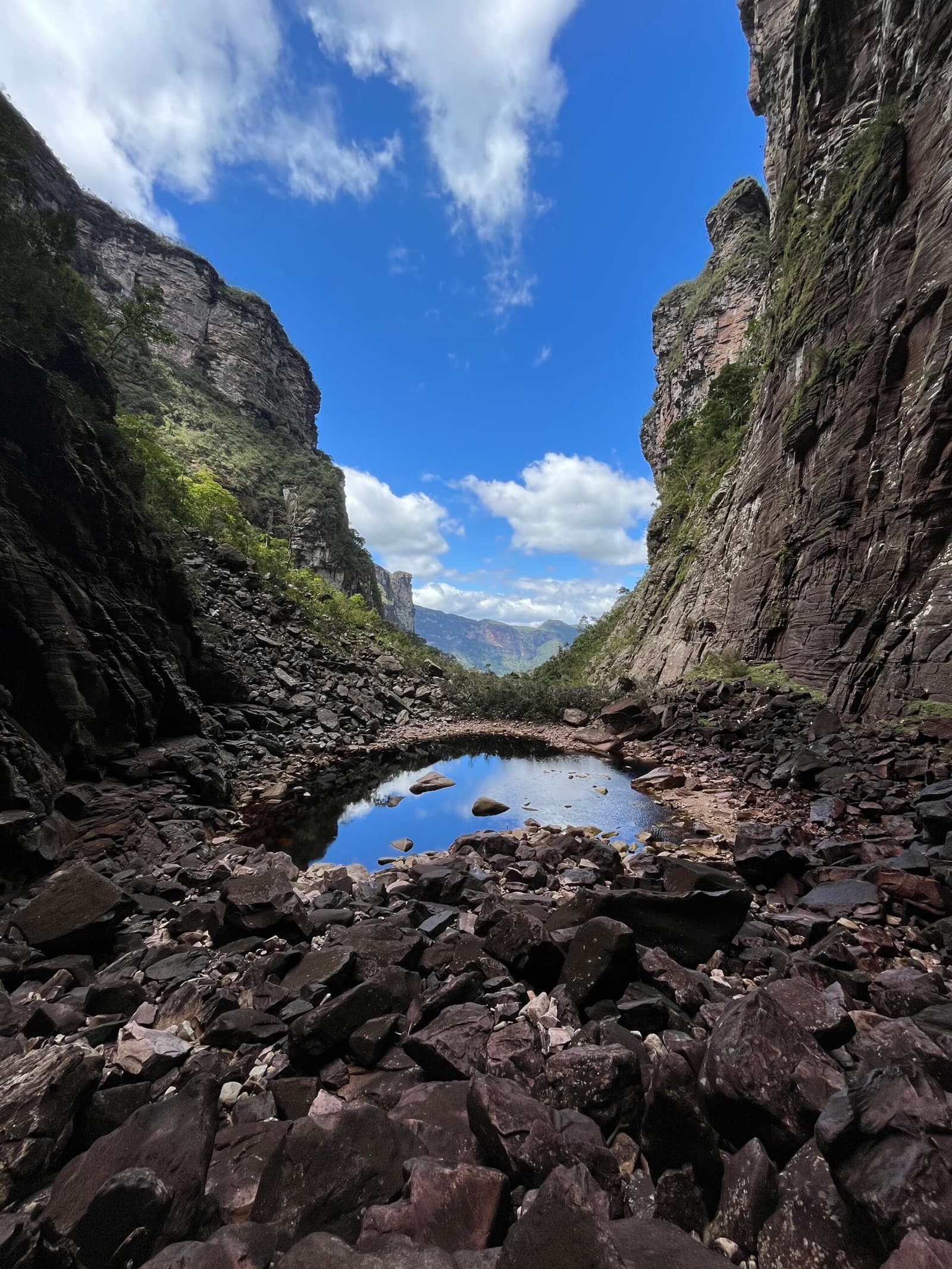 A pocket of sky, caught in stone and held still for you. Photo: Lucas Neves A narrow canyon on the Chapada Diamantina trek with towering sandstone walls flanking a still rock pool that mirrors the blue sky above, surrounded by dark boulders and opening to distant green mountains