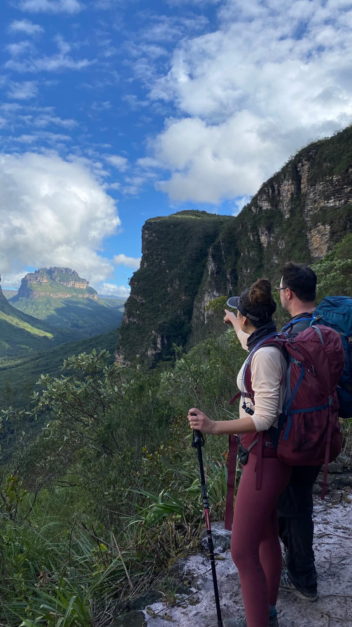 One hand points, and suddenly the whole valley makes sense. Photo: Lucas Neves Two hikers with backpacks and trekking poles pause at a rocky ledge on a Chapada Diamantina trek, one pointing toward the sweeping canyon valley and flat-topped sandstone mesas stretching under a partly cloudy sky