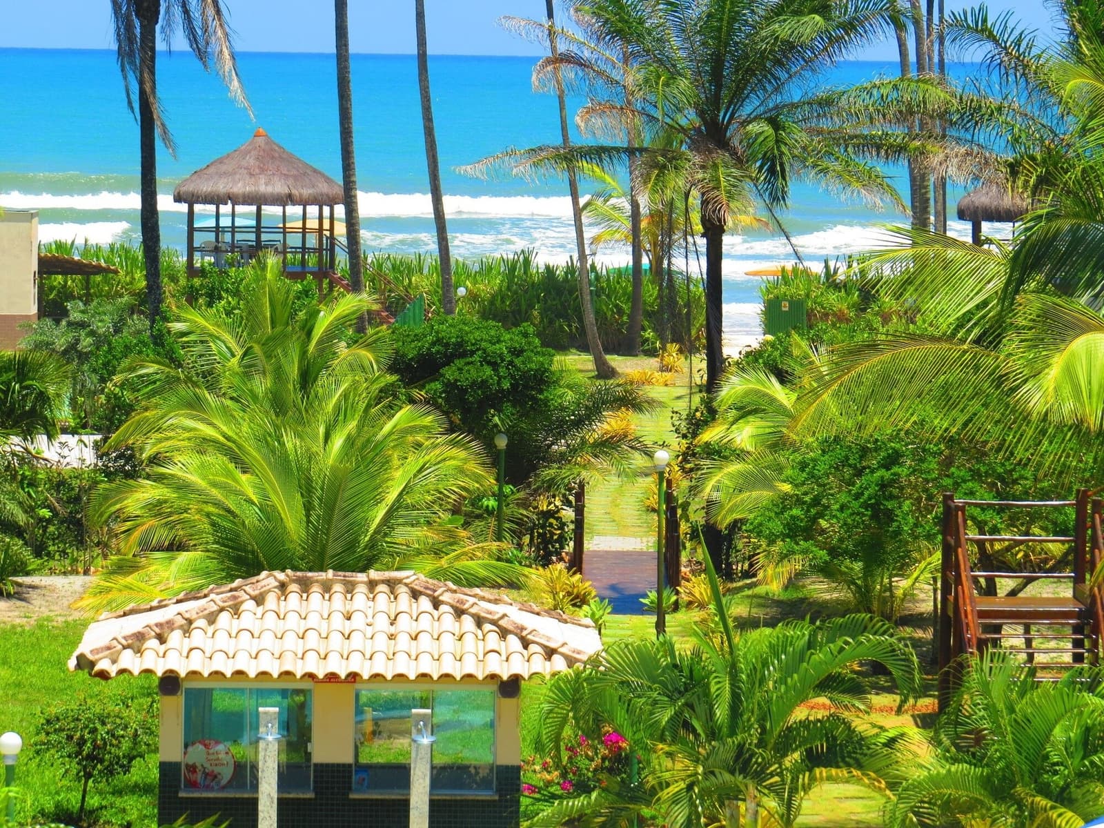 Your hotel garden is basically trying to upstage the ocean. It doesn't quite manage it. Lush tropical garden with palm trees and a tiled-roof kiosk leading via a brick path to a thatched beach gazebo and the Atlantic Ocean, a comfortable base for a cycling Bahia itinerary
