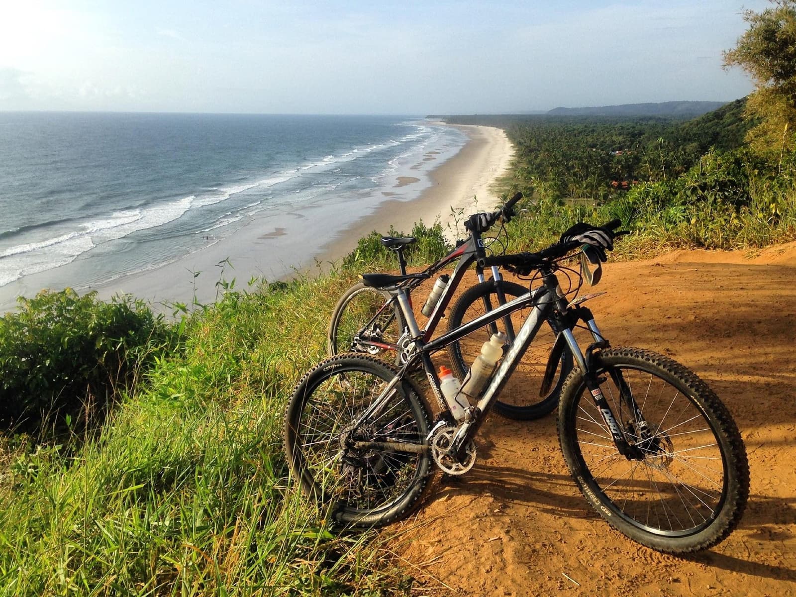 Two bikes, one view, and a beach that just keeps going. Two mountain bikes parked on a red dirt trail at a clifftop overlook during a cycling Bahia tour, with a long empty beach, Atlantic surf, and dense coastal forest stretching to the horizon below