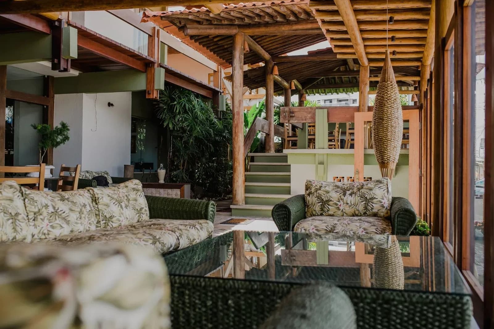 Rest well tonight, the road back to the saddle starts early. Open-air lounge with wicker sofas and floral cushions under a rustic log-beam roof, tropical plants and a staircase visible in the background at a guesthouse on the cycling Bahia Cocoa Route
