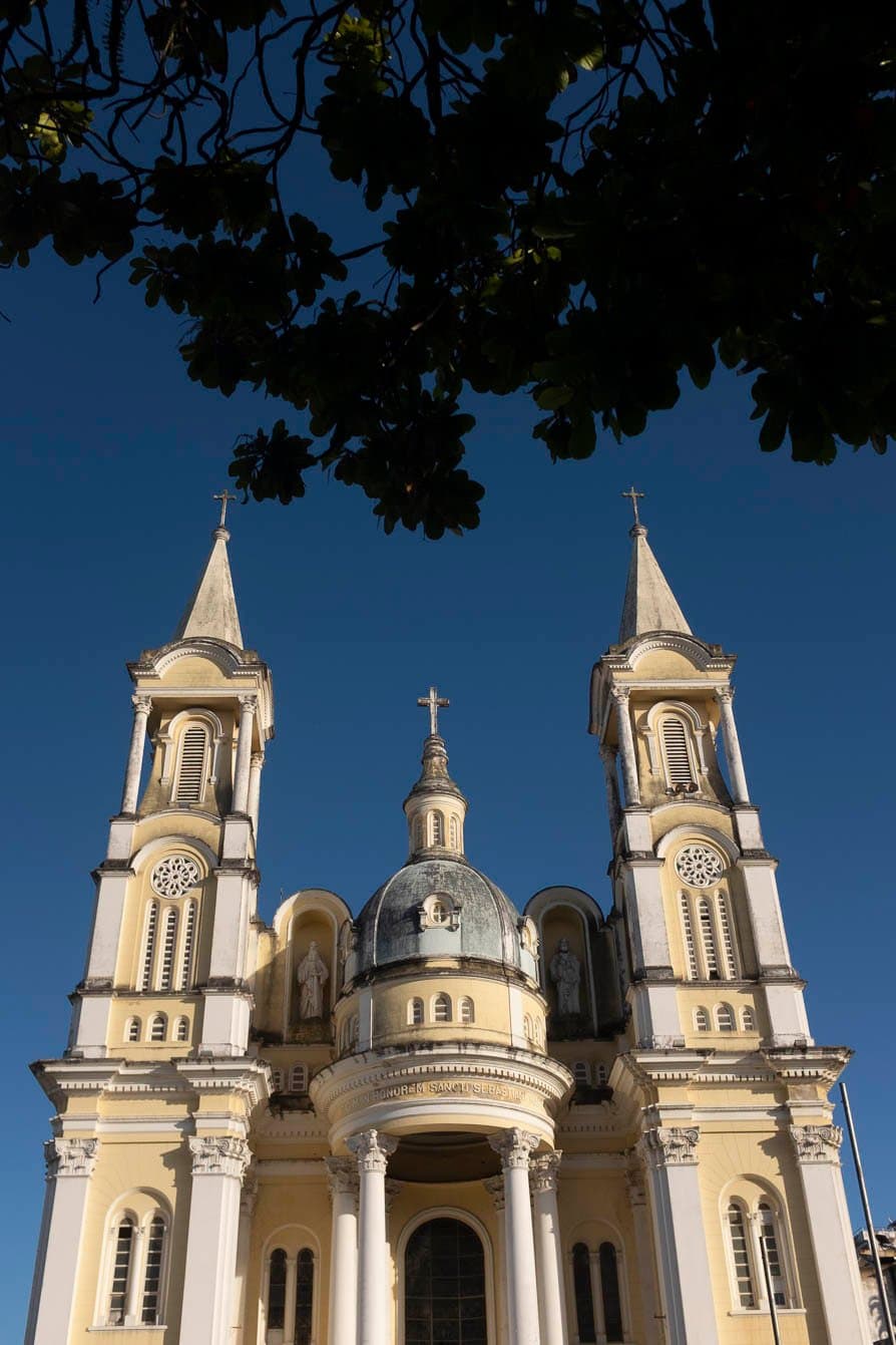 Ilhéus has a way of reminding you that history and sunshine arrive at the same time. Low-angle view of a pale yellow and white neoclassical church with twin bell towers, a central dome topped with a cross, and the inscription 'In Honorem Sancti Sebastiani' on its facade, framed by dark tree branches against a deep blue sky in Ilhéus, Bahia — the starting city of this cycling Bahia experience.