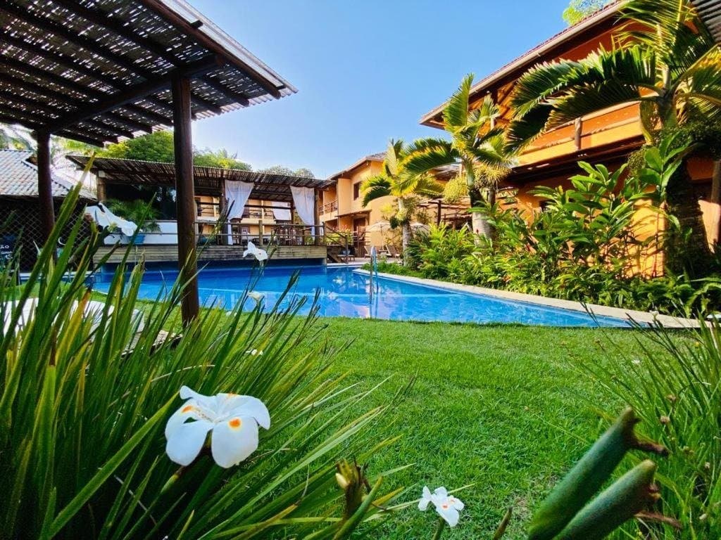Pedal hard, then land somewhere this green. Lush tropical garden with white flowers in the foreground, a blue swimming pool, and a warm-toned two-storey guesthouse under a clear sky on the cycling Bahia Cocoa Route in Itacaré