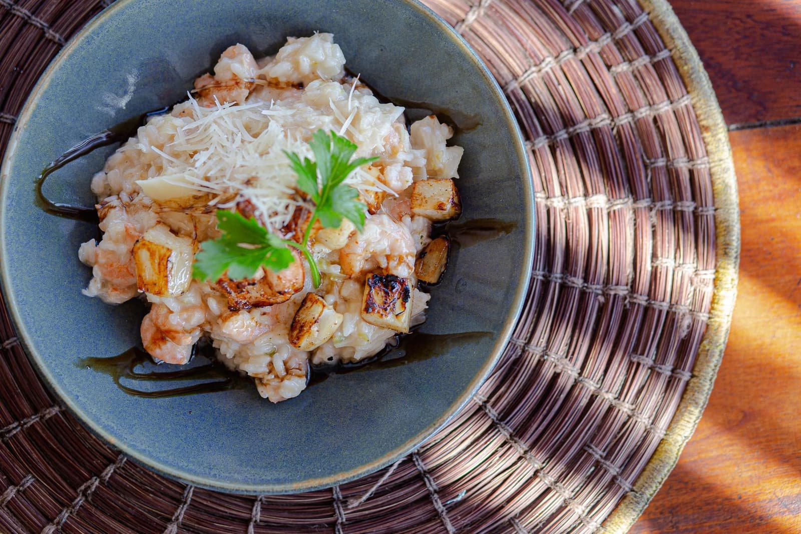 Legs rested, appetite earned, bowl full. Dinner sorted. A ceramic bowl of creamy shrimp risotto with charred root vegetable chunks, grated cheese, a fresh herb garnish and a balsamic drizzle, served on a woven rattan placemat on a wooden table — part of the dining experience on a cycling Bahia tour